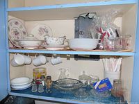 Full view of kitchen cupboard shelf containing plates, bowls, mugs, cutlery, and glassware.