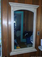 Photo of a large wall mirror with a cream-colored wooden frame hung on a brown wood-paneled wall, reflecting a room interior.