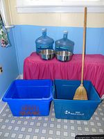 Two blue plastic recycling bins, two stainless steel bowls, whisk broom with wooden handle, and two large blue water jugs on pink tablecloth in corner with blue walls.