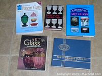 Five reference books on a tiled floor, showing covers with titles on pattern glass, American and Canadian goblets, American art glass, art glass price guide, and Cambridge Glass Co.