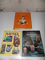 Three collector reference books related to Majolica, Weller pottery, and half dolls displayed on a table.