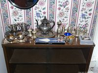 Overall display of the Indian silverplate set and assorted items on a wooden surface in front of floral wallpaper.