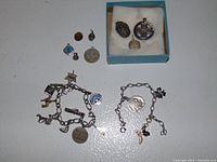 Two silver-tone charm bracelets laid out flat with multiple attached charms along with additional loose charms placed above one bracelet, some charms in a blue box.