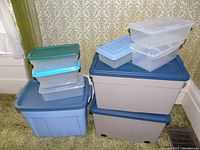 Photo showing multiple plastic storage totes and containers of various sizes and colors stacked and arranged, including beige totes with blue lids and smaller clear and colored containers.