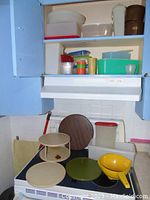 View of beige and wood-toned lazy susans on stove top with assorted plastic bowls and trays inside cabinets above stove.