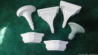 Six white decorative wall pieces including three wall sconces and three corner shelves laid out on green fabric background. Visible variations in shape and detail among sconces and shelves.