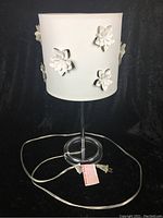Photo of the lamp showing the white shade with 3D floral cutouts and clear base on a black background, lamp unlit.