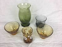 Six pieces of assorted glassware on a white background including a green ribbed vase, three amber glass bowls, one amber floral tumbler, and one smoky glass.