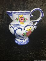 Front and side view of white ceramic pitcher with blue detailing and colorful floral hand-painted design, showing shape and artwork