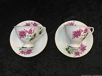 Pair of matching porcelain teacups with saucers featuring fuchsia flower design on a black background.