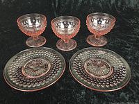 Front view of three pink hobnail footed dessert cups and two hobnail plates laid flat on black background.