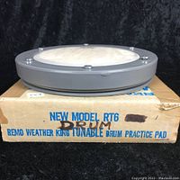 Side view of the round grey practice drum pad sitting on original cardboard box. The box is labeled NEW MODEL RT6 REMO WEATHER KING TUNABLE DRUM PRACTICE PAD with some handwritten markings.