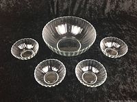 Photo showing one large and four small glass bowls arranged on a black surface, all clear with scalloped edges.