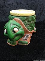 Front view of the turtle-shaped plant pot, showing the large green head with sculpted eyes and the textured green shell with a cream circular top.