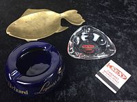 Three vintage ashtrays displayed together including brass fish-shaped, blue ceramic, and clear glass ashtray with label.