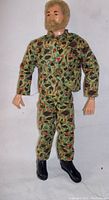 Front view of the 1964 GI Joe action figure showing fuzzy blonde hair and beard, wearing multi-color camouflage jacket and pants, and black boots.