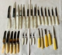 Top-down view of cutting utensils showing two rows of knives with mother of pearl and opaque/faux bone handles, four forks with Bakelite handles, several small two-prong forks, and specialty cutting tools with yellow Bakelite handles arranged on a white cloth.