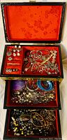 Open jewelry box showing three tiers filled with assorted vintage jewelry on red velvet lining with floral design inside lid
