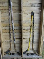 Four hockey sticks and four hockey pucks arranged vertically against a wood wall, showing condition and brands.