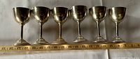 Front view showing the set of six silver plated wine goblets, aligned side by side with ruler measuring approximately 5 inches in height and 2 inches in diameter at the rim.
