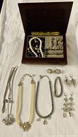 Opened handcrafted wooden jewelry box showing pearl necklace, brown and beige beaded necklace, and various rings and earrings arranged in compartments.