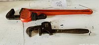 Photo of large 14 inch pipe wrench with orange handle and smaller 10 inch pipe wrench laid flat on a surface.