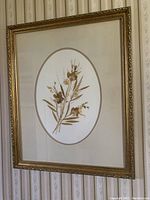 Close-up photo of one framed pressed flower artwork featuring an oval mat with detailed gold frame and intricate dried flowers and leaves.