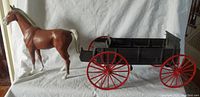 Brown horse with white mane and tail standing beside a black wagon with red wheels on a white backdrop.