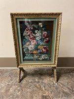 Gold ornate folding picture frame standing upright with floral needlepoint artwork inside.