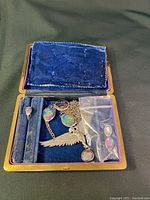 Open blue velvet-lined jewelry case showing necklace with wing pendant and oval gemstones, ring, and earrings; earrings in a plastic bag, one missing hook
