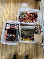 Three plastic bins containing assorted tools and hardware on wood floor, including hand saws, coiled extension cords, battery-operated air compressor, and other tools.
