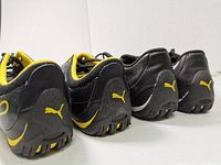 Rear view of two pairs of Puma soccer shoes showing the heel logos and cleats, black and yellow and all black color schemes.