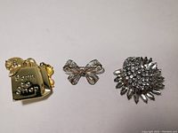 Full view of the three brooches on a plain surface showing the Born to Shop bag, filigree bow, and rhinestone floral brooch from left to right.