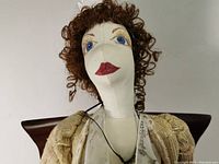 Close-up of cloth doll's face showing painted blue eyes, red lips, and curly brown hair with necklace.