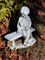 Plaster statue of young boy sitting on bench with two dogs, surrounded by garden foliage, photograph showing front and side views