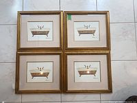 Four artist prints arranged in a 2x2 grid on floor, each print framed in gold colored frame with cream matte, depicts clawfoot bathtubs with gold detailing.