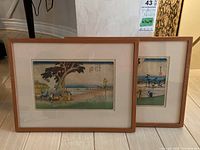 Two framed Asian prints, one behind the other, showing coastal scenery and people by trees, matted and framed in matching light wood frames.