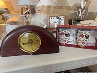 Photo showing vintage wooden weather station with brass barometer, two floral butterfly patterned teacups in red gift box, clear glass plates, glassware, and white string lights on white table.