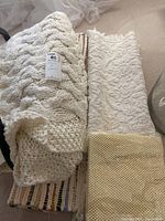 Photo showing chunky knit cream throw blanket, cream textured crochet-like blanket, woven multicolor floor mat, and beige waffle-patterned floor mat stacked together.