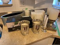 Photo of various candle holders including two metal pillar holders with white candles, boxed Raquel Breane candle holders, iridescent glass holders, and vases.