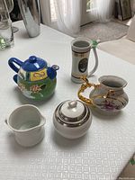 Photo of colorful teapot with abstract and floral design, white ceramic creamer, white sugar bowl with lid and silver trim, white ceramic pitcher, and porcelain vase with gold crane handle and floral painting.