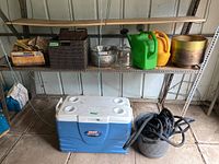 Wide shot of all items including Coleman cooler, synthetic baskets, metal plant pot, watering cans, glass terrarium and bowls, box of Miracle Mulch, work gloves, and garden hose in resin pot on metal shelving and floor