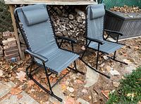 Two folding outdoor rocking lawn chairs with grey mesh fabric and black frames positioned outdoors on stone and leaf-covered ground near a woodpile. Each has headrests and armrests, one with a missing plastic cap on frame.