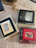 Stack of assorted picture frames and photo albums on a white surface, showing various frame styles and sizes including black, cream, and red leather album.