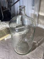 Clear large glass jug standing upright on carpeted floor, showing full height and cylindrical shape with narrow neck opening.