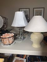 Three lamps photographed on a table including a Himalayan salt lamp in a black basket, a layered glass lamp with white shade, and a beige ceramic lamp with large shade.