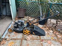 Full view of black Poulan Pro lawn mower with yellow wheels and grass catcher bag on paved outdoor surface showing use and dirt.