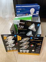 Box containing unopened and opened packages of Luminus LED bulbs and Gardeners Eden solar patio lights on floor near window.