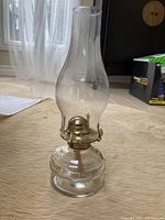 Clear glass oil lamp with a round base, brass burner, and tall chimney on a beige carpet.