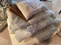 Stack of white pillows, brown knit throw blanket, and white duvet cover piled on beige surface.
